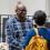 A man in a checked shirt talking to another man in a blue jumper with a yellow backpack