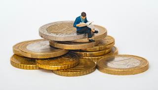 A small doll reading  a book whilst sitting on top of a pile of coins