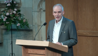 Roland Bleiker at a lectern in a grey suit with a white shirt delivering a keynote speech