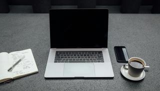 An open computer with a coffee and phone on the right hand side and an open notebook with a pen and writing inside on the left