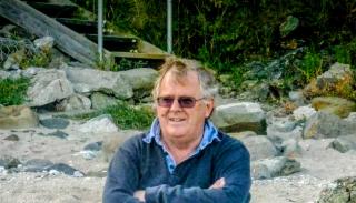 John Groom sat on a rock at the beach wearing a blue sweater