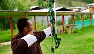 A man shooting an arrow out of a bow in a green field with a covered walkway in the background