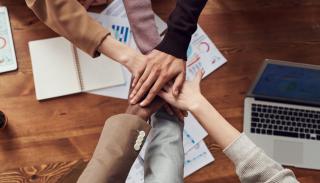 A group of people of different ethnicities joining hands over a table.