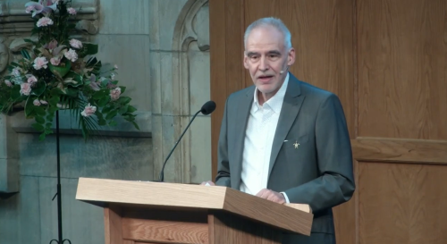Roland Bleiker at a lectern in a grey suit with a white shirt delivering a keynote speech