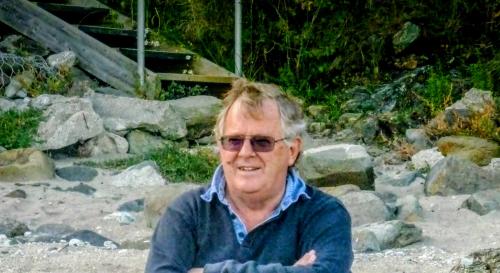 John Groom sat on a rock at the beach wearing a blue sweater