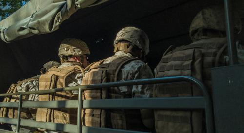Soldiers dressed in camouflage lined up with their backs to the camera seated in a military truck with a green awning