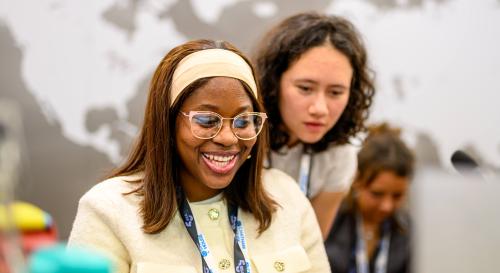 A woman with glasses and a beige shirt and headband smiling whilst using a laptop, with another woman reading over her shoulder